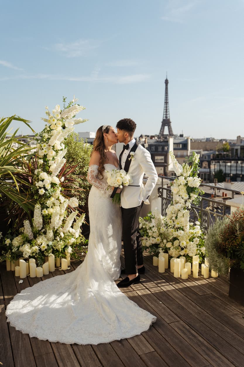 Bride and groom kiss on a flower-lined rooftop terrace with the Eiffel Tower in the background