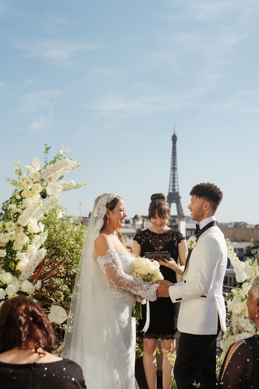 Bride and groom hold hands before an officiant with guests, the Eiffel Tower rising behind them in bright sunlight