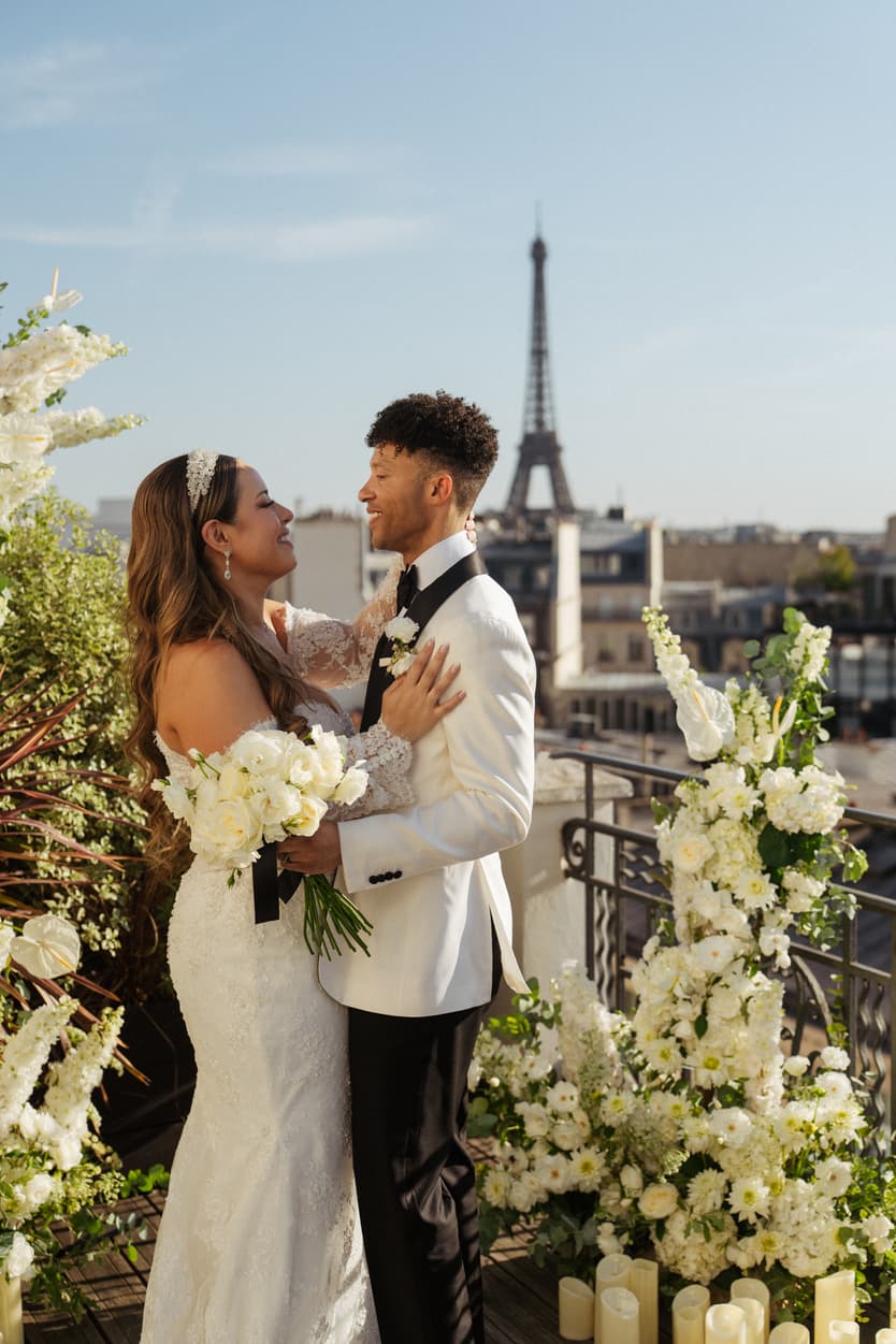 Bride and groom embrace on a flower-lined terrace with the Eiffel Tower behind them in warm sunlight