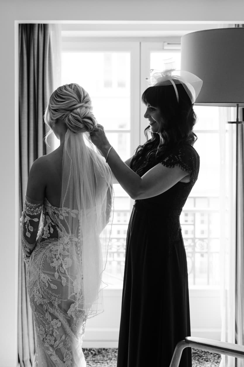 Bride in lace veil gets her hair adjusted by a woman in a black dress beside a bright hotel window