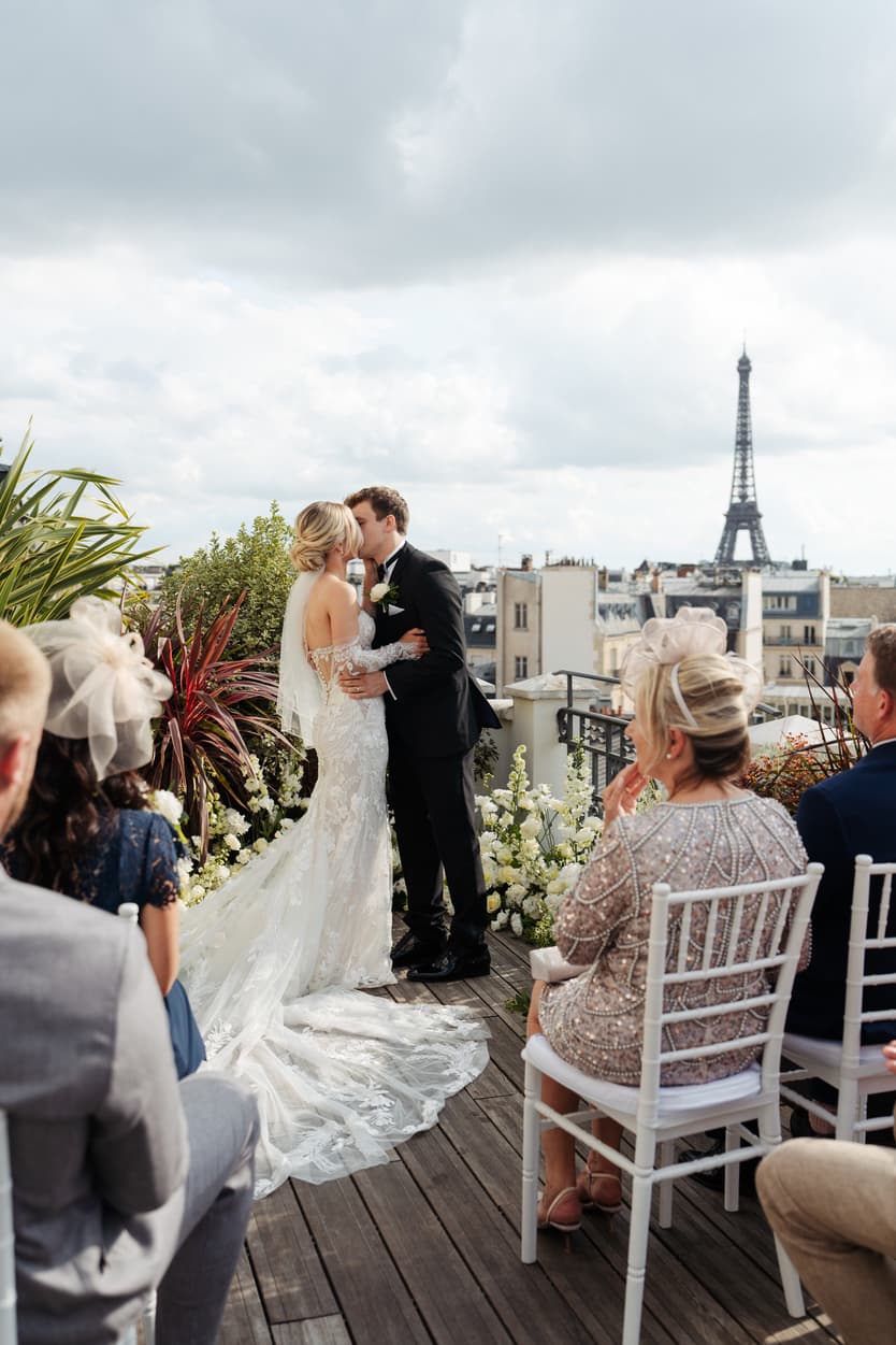 Bride and groom kiss on a rooftop terrace with guests seated and the Eiffel Tower beyond under soft light