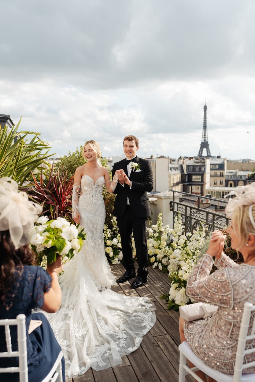 A bride and groom laughing before their guests on the flowered rooftop terrace of Hotel Marignan, the Eiffel Tower behind