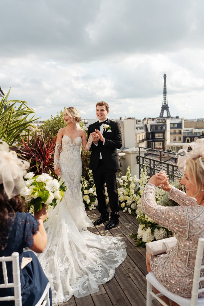 Bride and groom stand with guests on a rooftop terrace, Eiffel Tower in view beyond white flowers