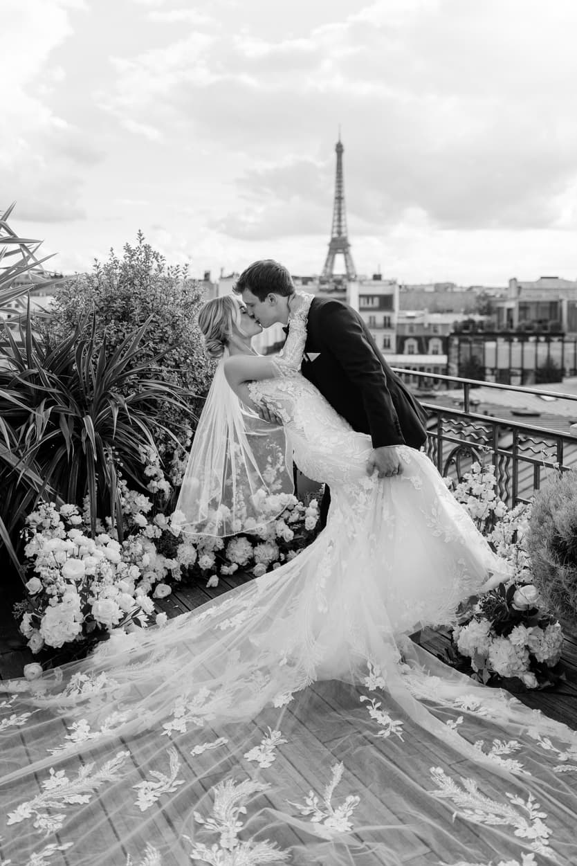 Bride and groom kiss on a flower-lined rooftop terrace with the Eiffel Tower behind them