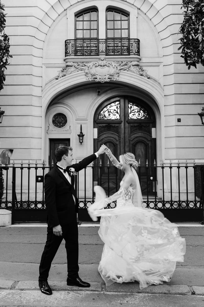 Bride and groom twirl on a Paris street before an ornate townhouse gate in soft black and white light