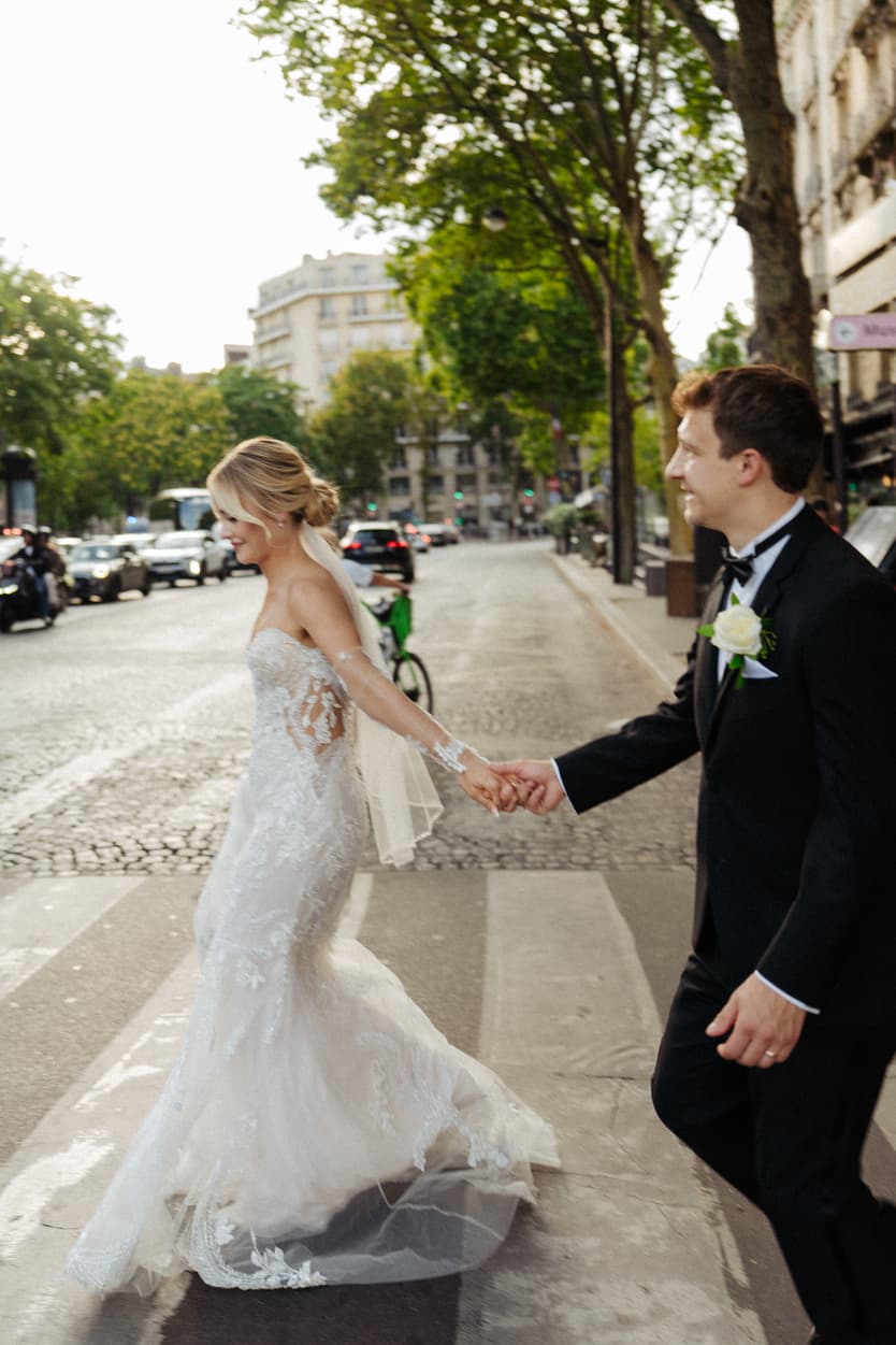 Bride and groom cross a Paris street at golden hour, hand in hand, her lace veil trailing behind