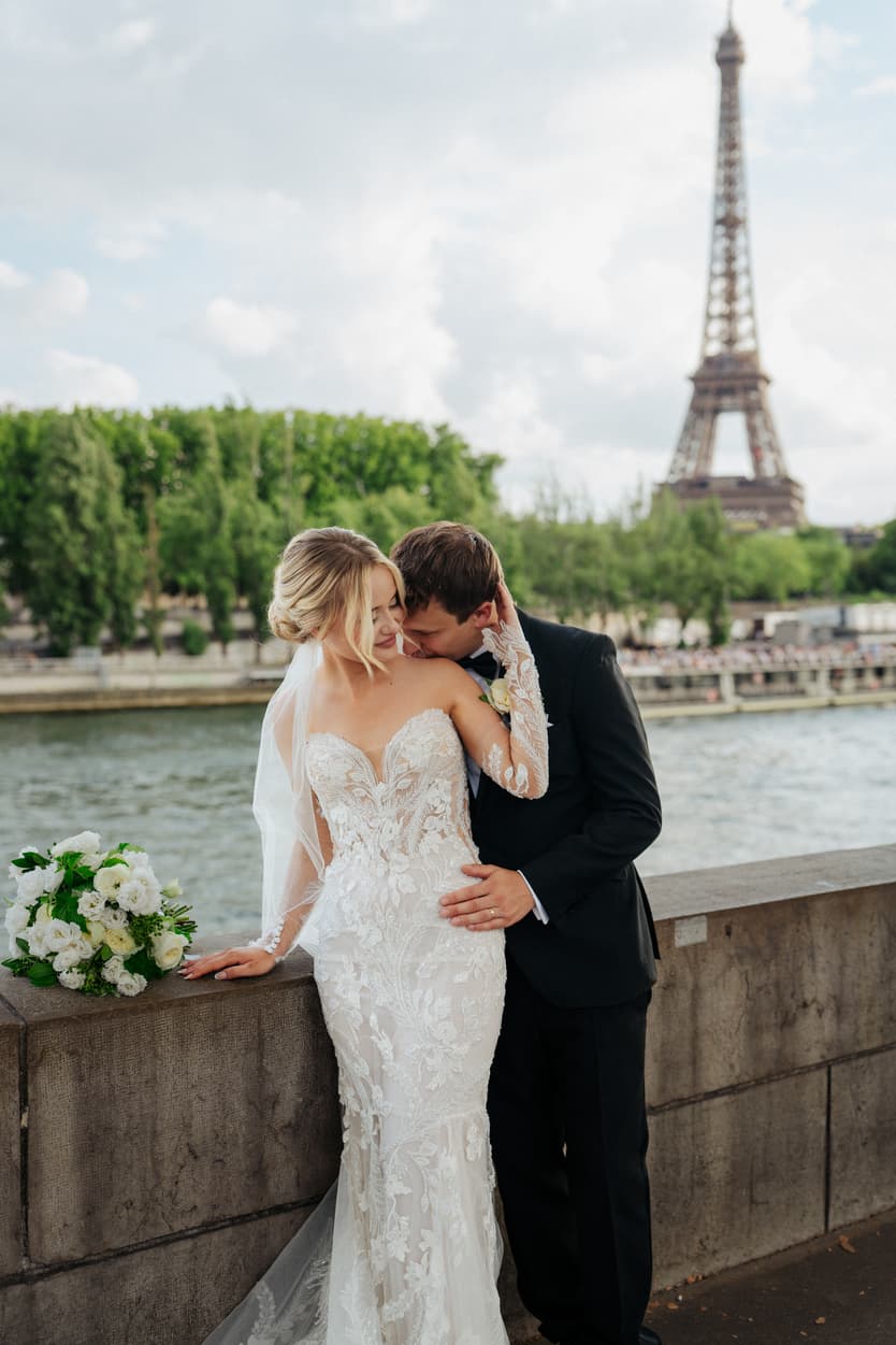 Bride and groom embrace by the Seine with the Eiffel Tower behind them in soft daylight, bouquet on the wall