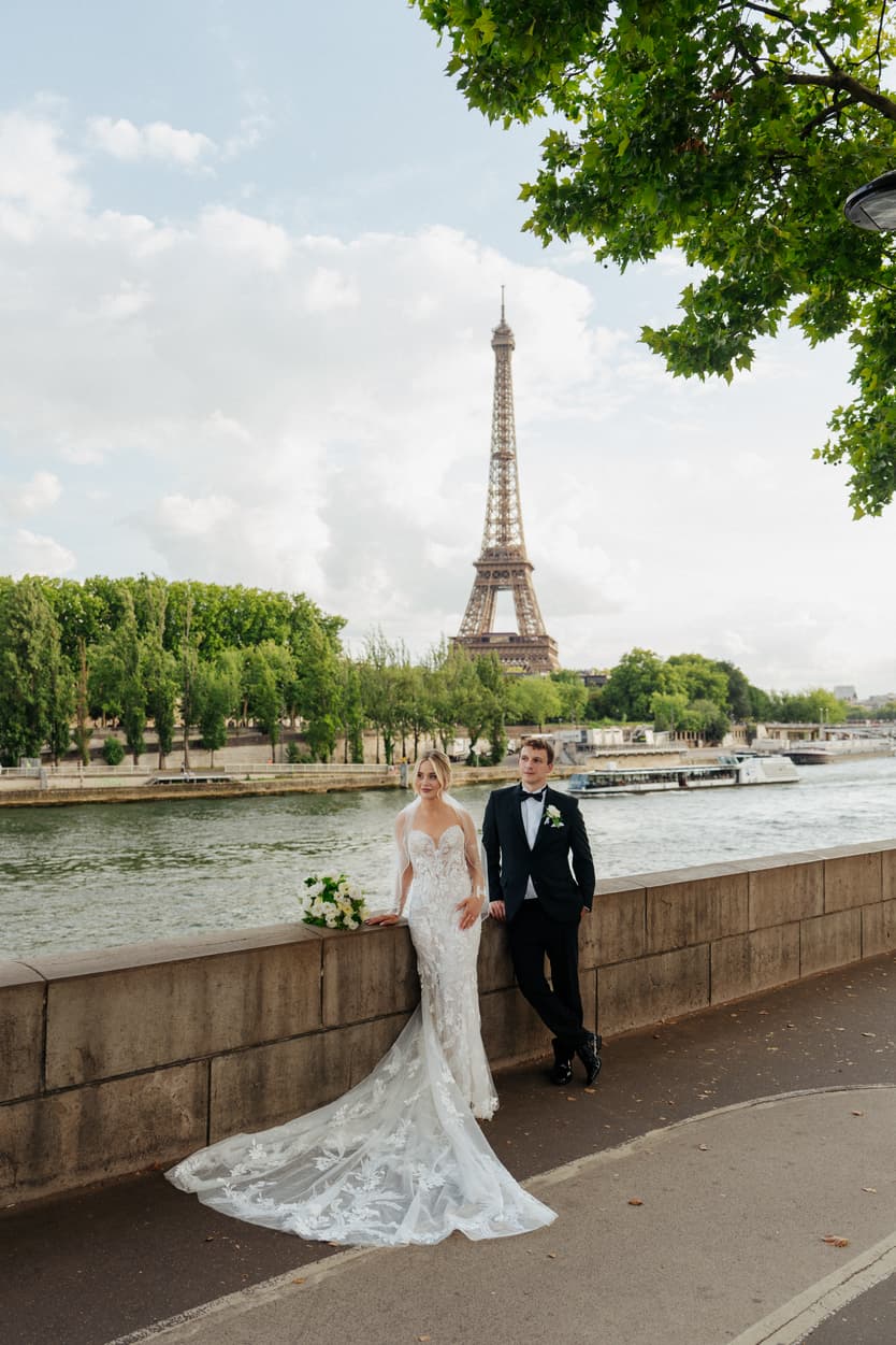 Bride and groom pose by the Seine with the Eiffel Tower behind them in soft daylight