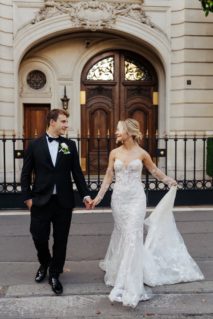Bride and groom hold hands on a Paris street before an ornate doorway in soft daylight