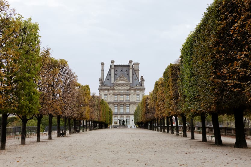 A long tree-lined garden path leads to a grand Paris building under soft daylight