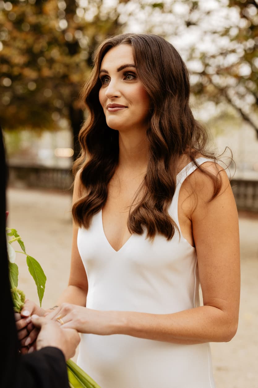 Bride in a white dress holds hands during an outdoor ceremony in soft light, with greenery nearby