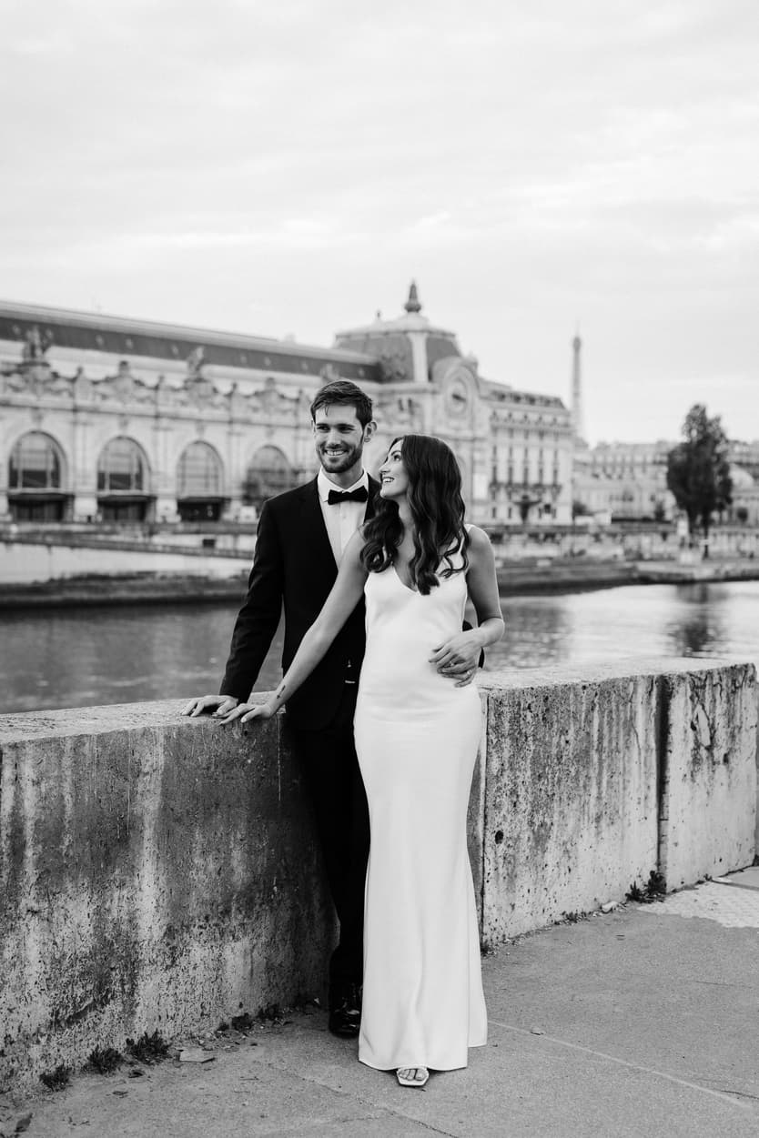 Bride and groom pose by the Seine, smiling beside a stone wall with the museum and Eiffel Tower beyond
