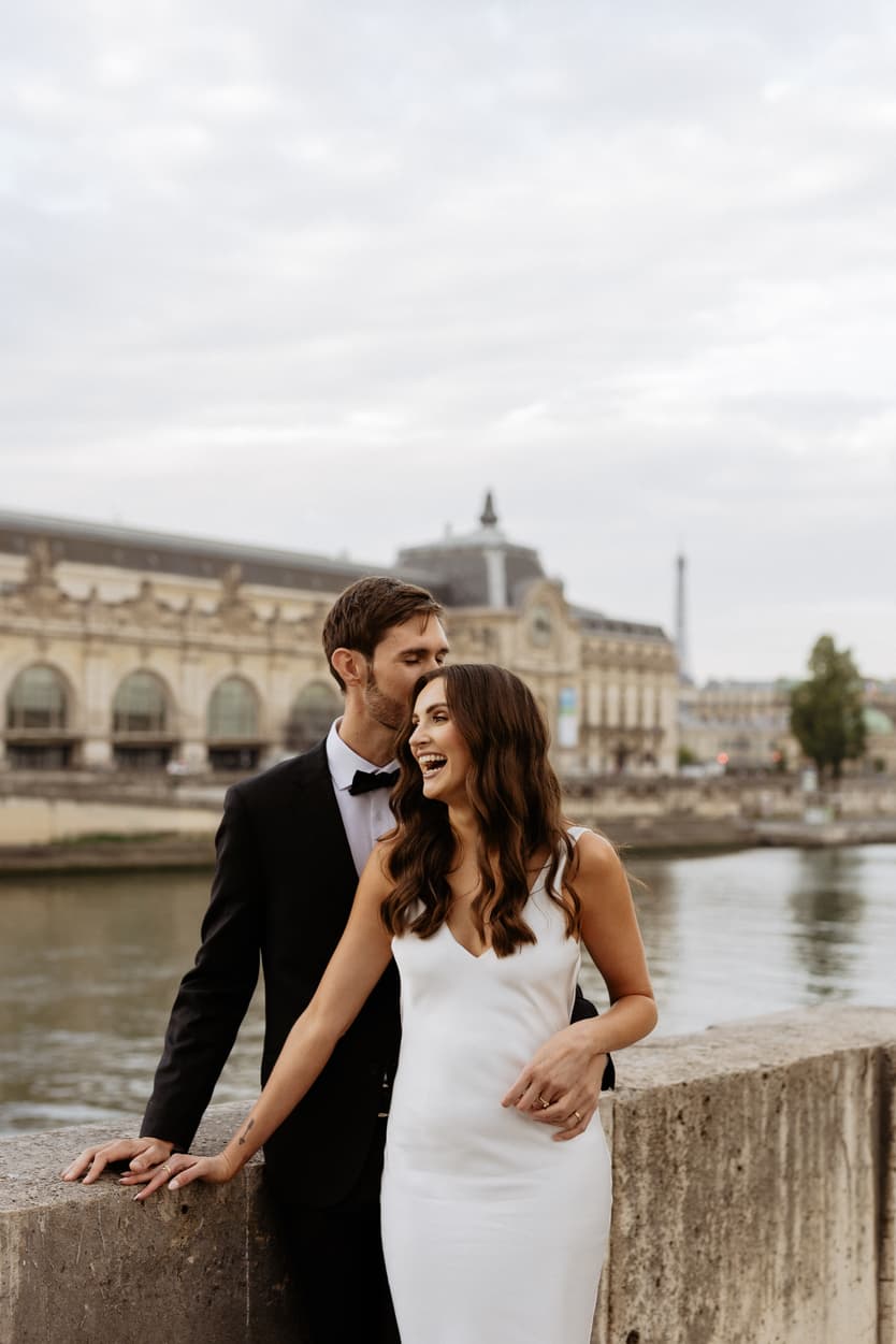 Bride and groom pose by the Seine, with a laughing kiss and a stone parapet in soft daylight