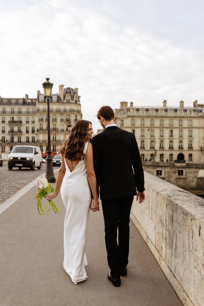 Bride and groom walk hand in hand along a Paris bridge, bouquet in her left hand, under soft gray light