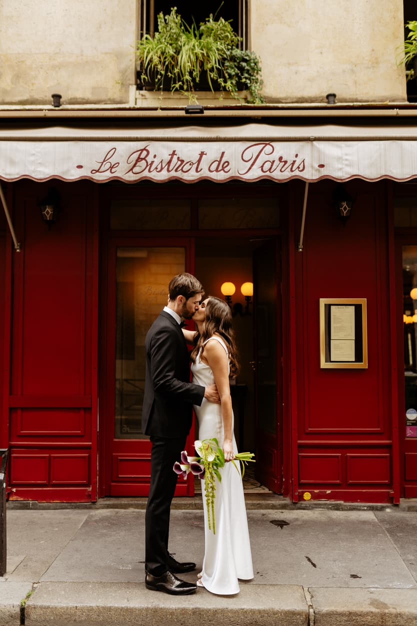 Bride and groom kiss outside Le Bistrot de Paris on a Paris street, her bouquet at her side