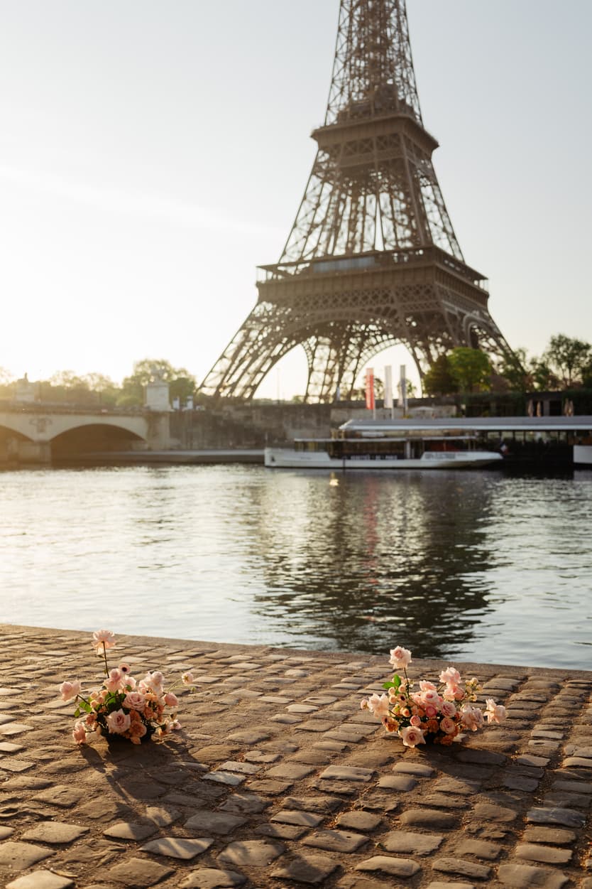 Bouquets rest on cobblestones by the Seine with the Eiffel Tower glowing in warm evening light