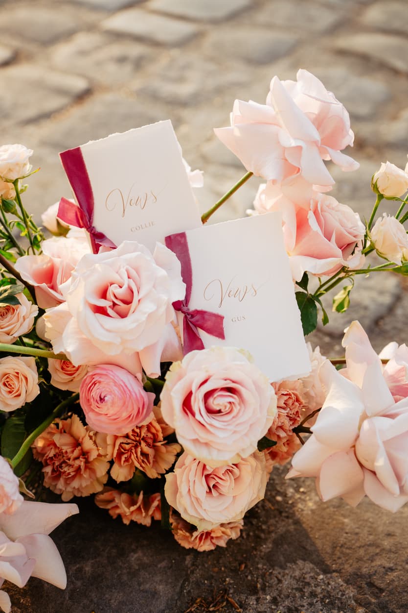 Blush roses and vow cards with magenta ribbons rest on sunlit Paris cobblestones