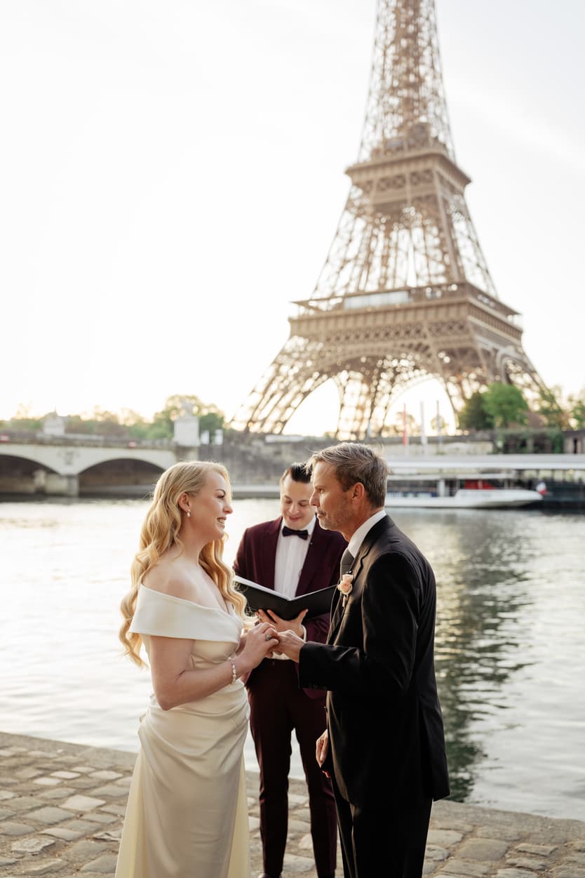 Bride and groom exchange rings by the Seine with the Eiffel Tower behind them as the officiant reads