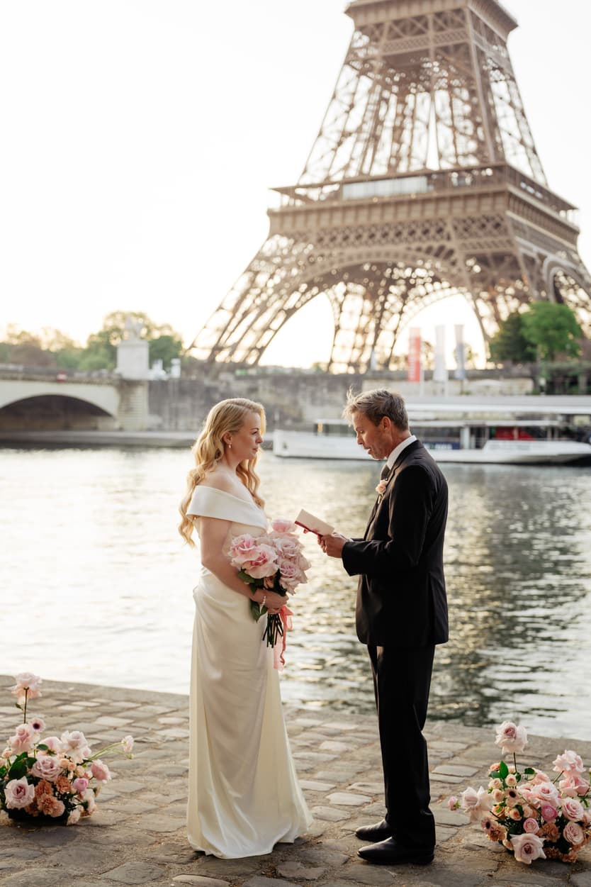 Bride and groom exchange vows beside the Seine, bouquet in hand, with the Eiffel Tower glowing behind them at sunset