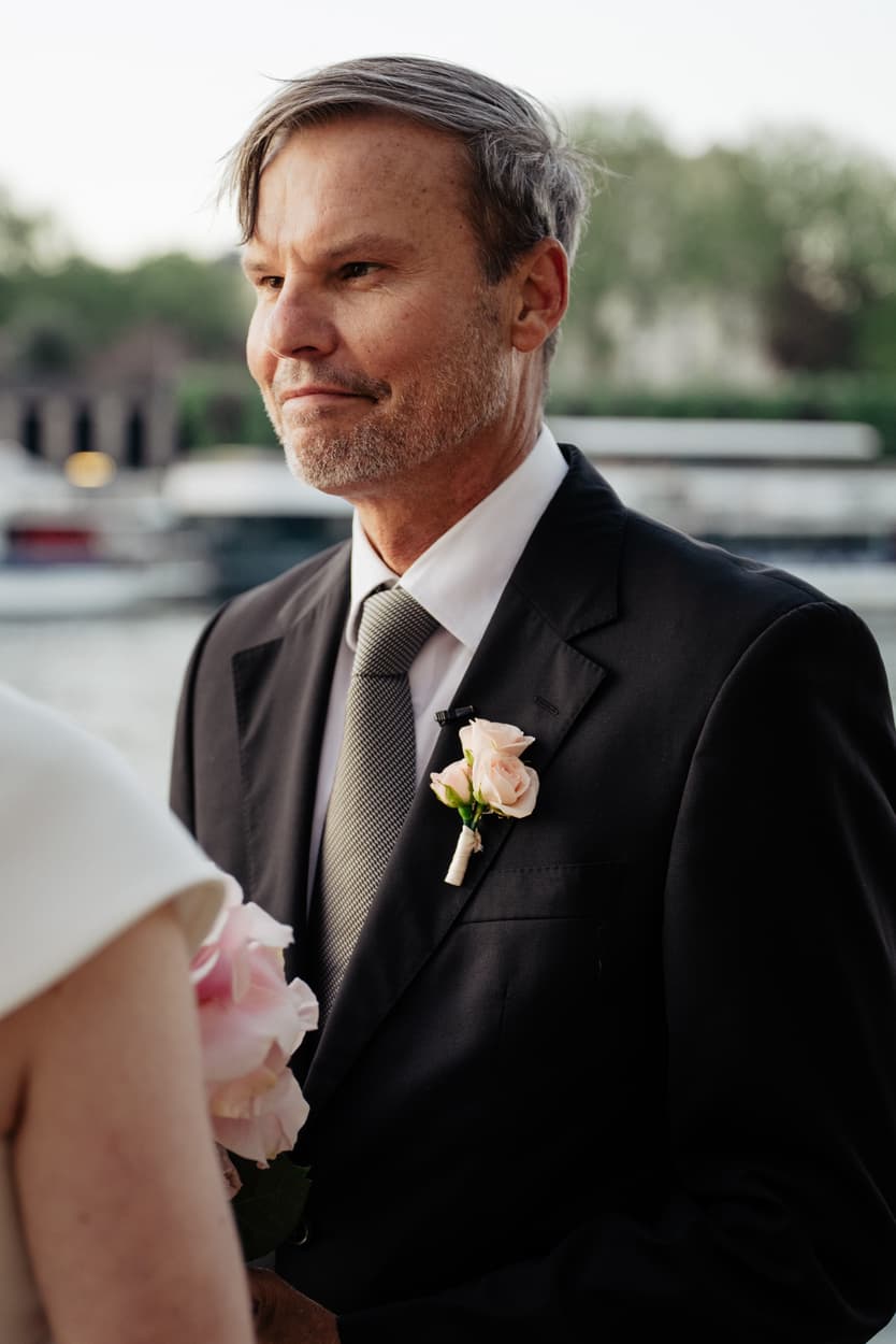 Groom in a black suit with boutonniere faces the bride on a Paris street in soft daylight