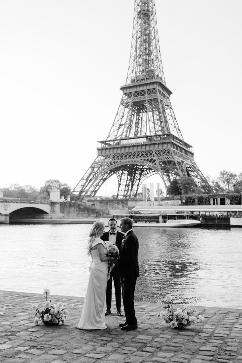 Bride and groom stand with an officiant by the Seine, framed by the Eiffel Tower and two floral arrangements