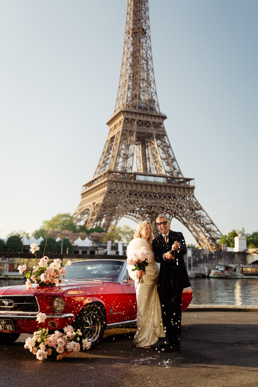 Bride and groom toast beside a red vintage car with flowers at the Eiffel Tower in warm evening light