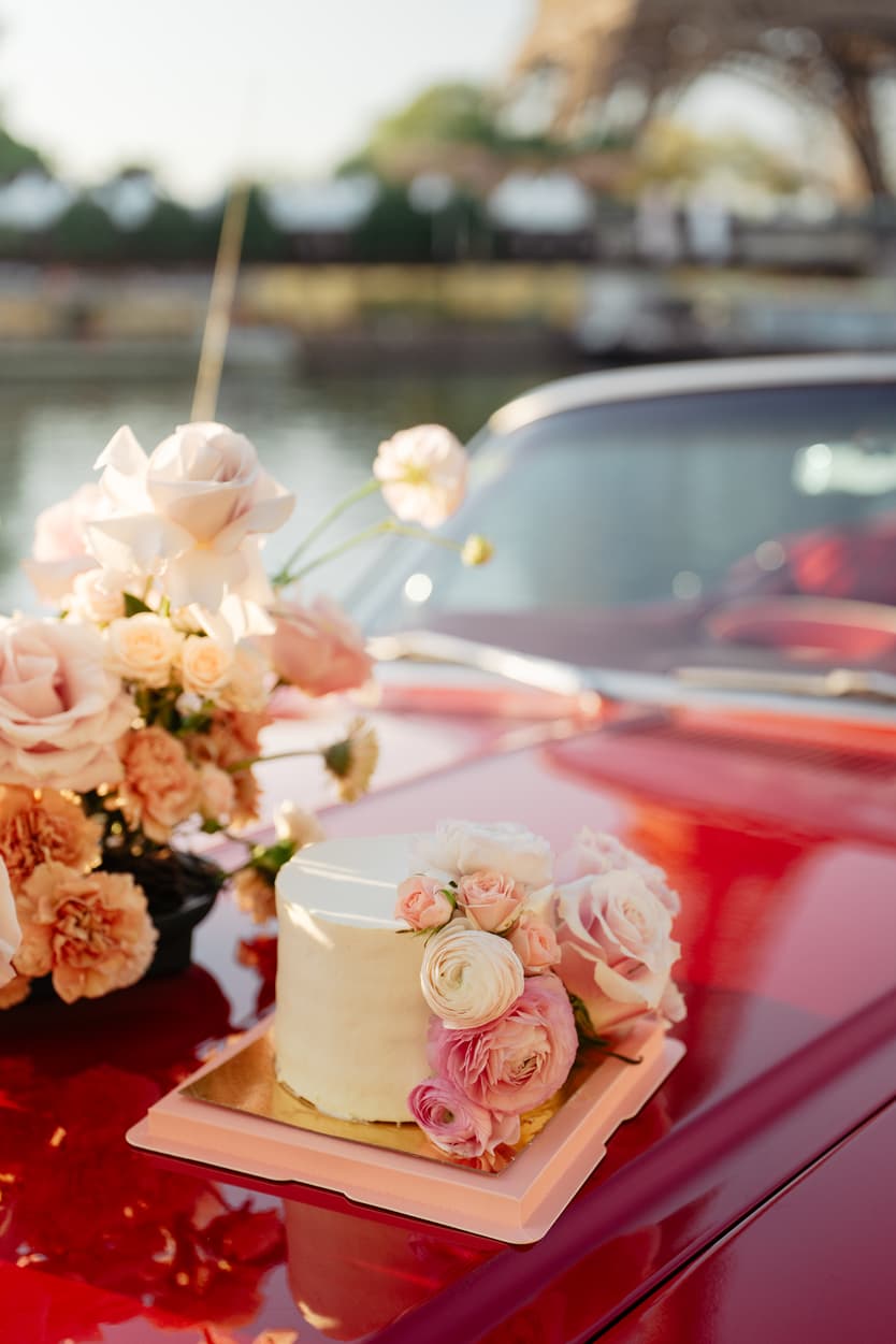Pink wedding cake and blush roses rest on a red car in warm waterfront light, with a blurred Paris landmark behind
