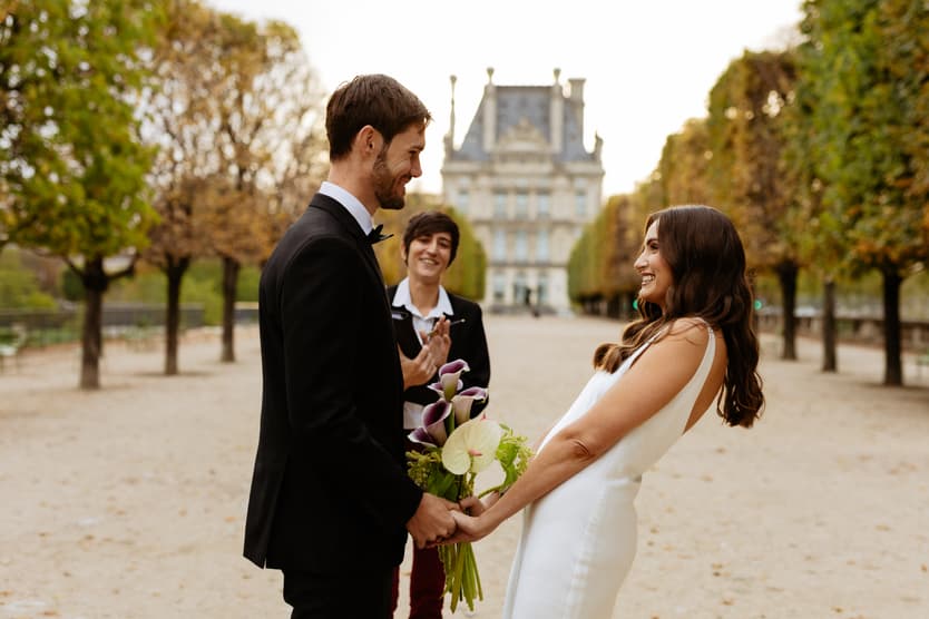 Bride and groom hold hands in a Paris garden as an officiant smiles behind them in soft daylight