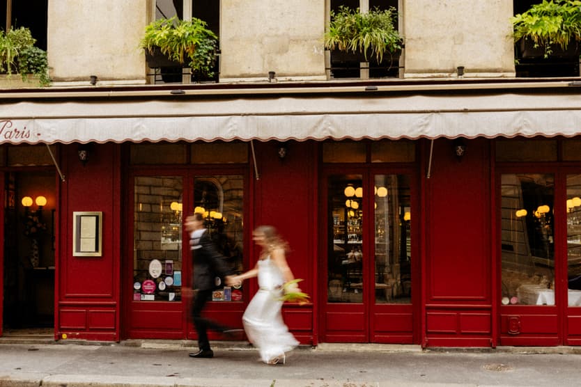 Bride and groom stride past a red Paris storefront, blurred in motion under warm window lights