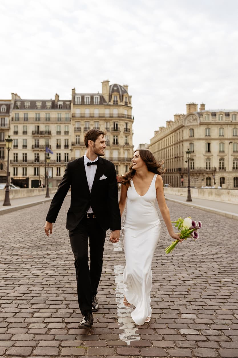 Bride and groom walk hand in hand on a Paris street, she carries purple tulips in soft daylight