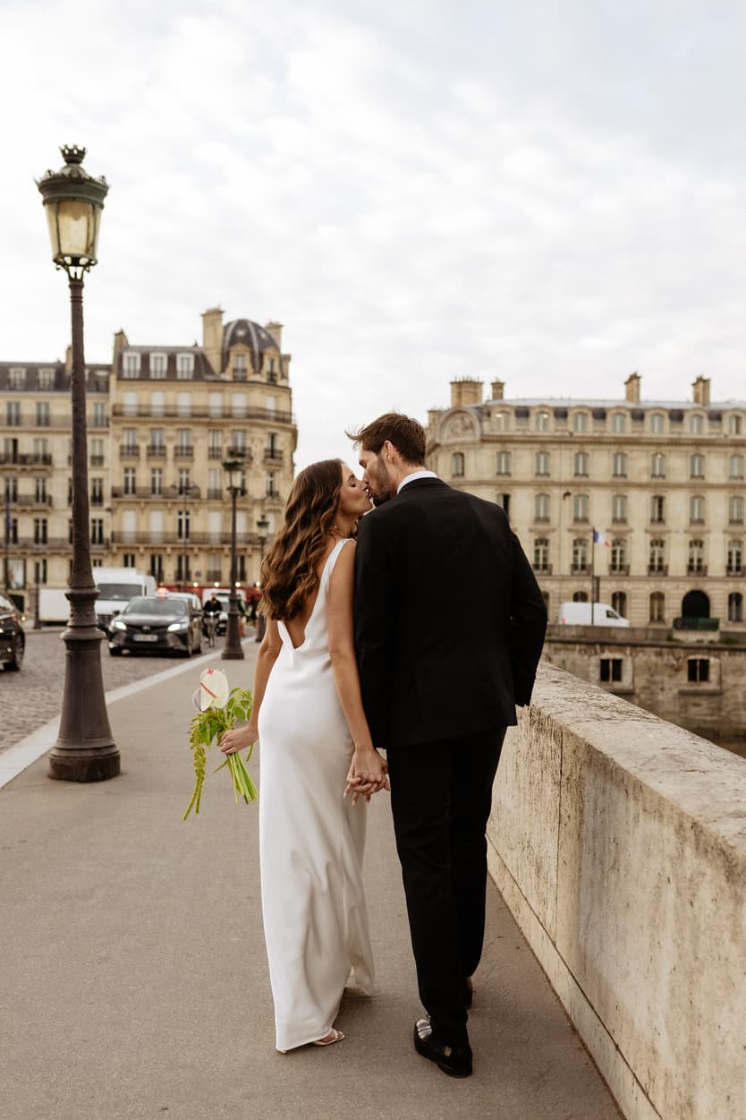 Bride and groom kiss on a Paris bridge at soft daylight, holding a bouquet and hand in hand