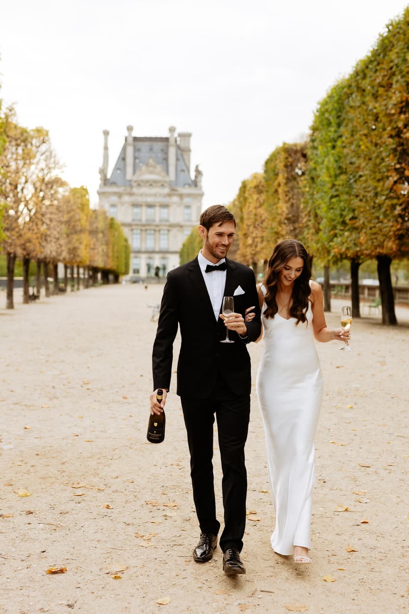 Bride and groom walk arm in arm through a Paris garden, carrying champagne under soft autumn light