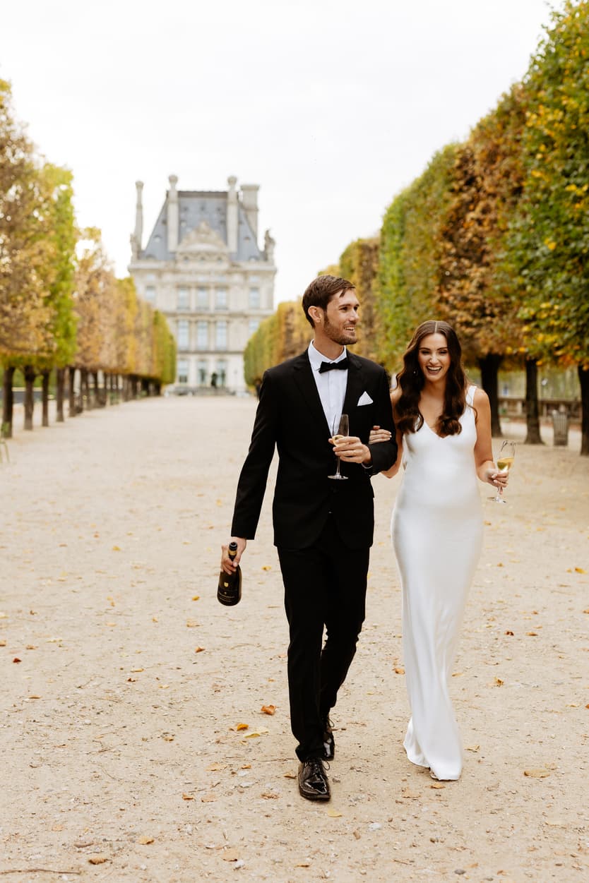 Bride and groom walk arm in arm through a Paris garden, holding champagne in soft autumn light