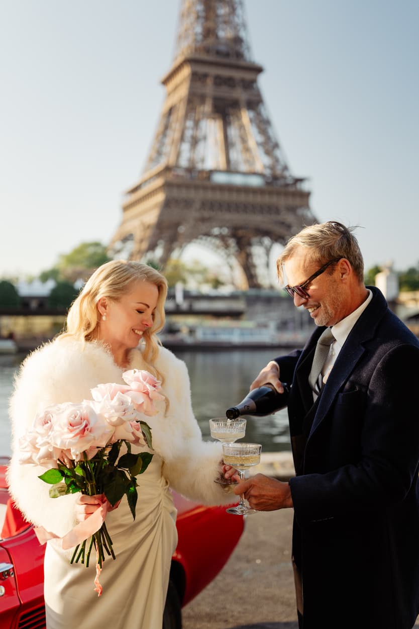 Bride and groom toast champagne by the Eiffel Tower, she holding pink roses in warm sunlight