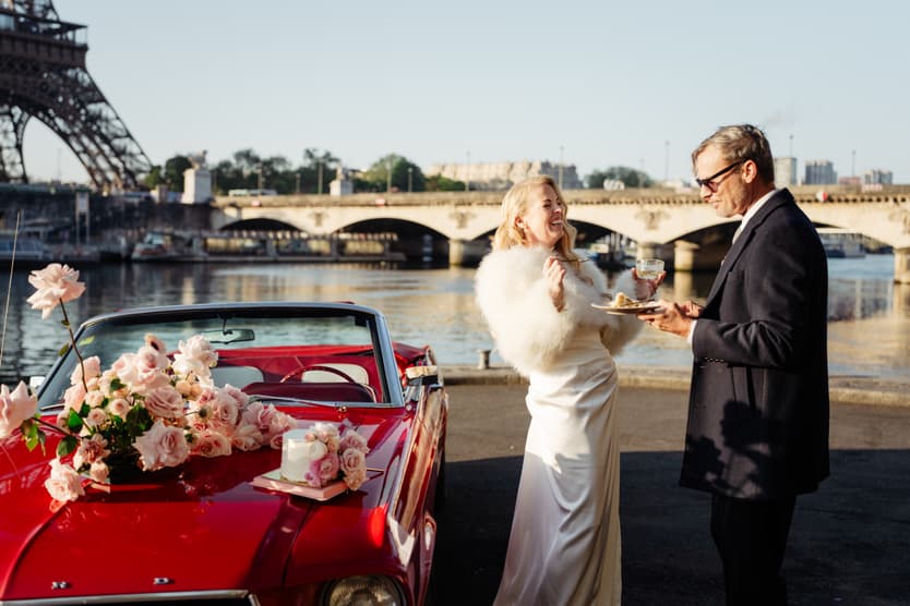Bride and groom toast beside a red flower-covered convertible by the Seine with the Eiffel Tower behind them