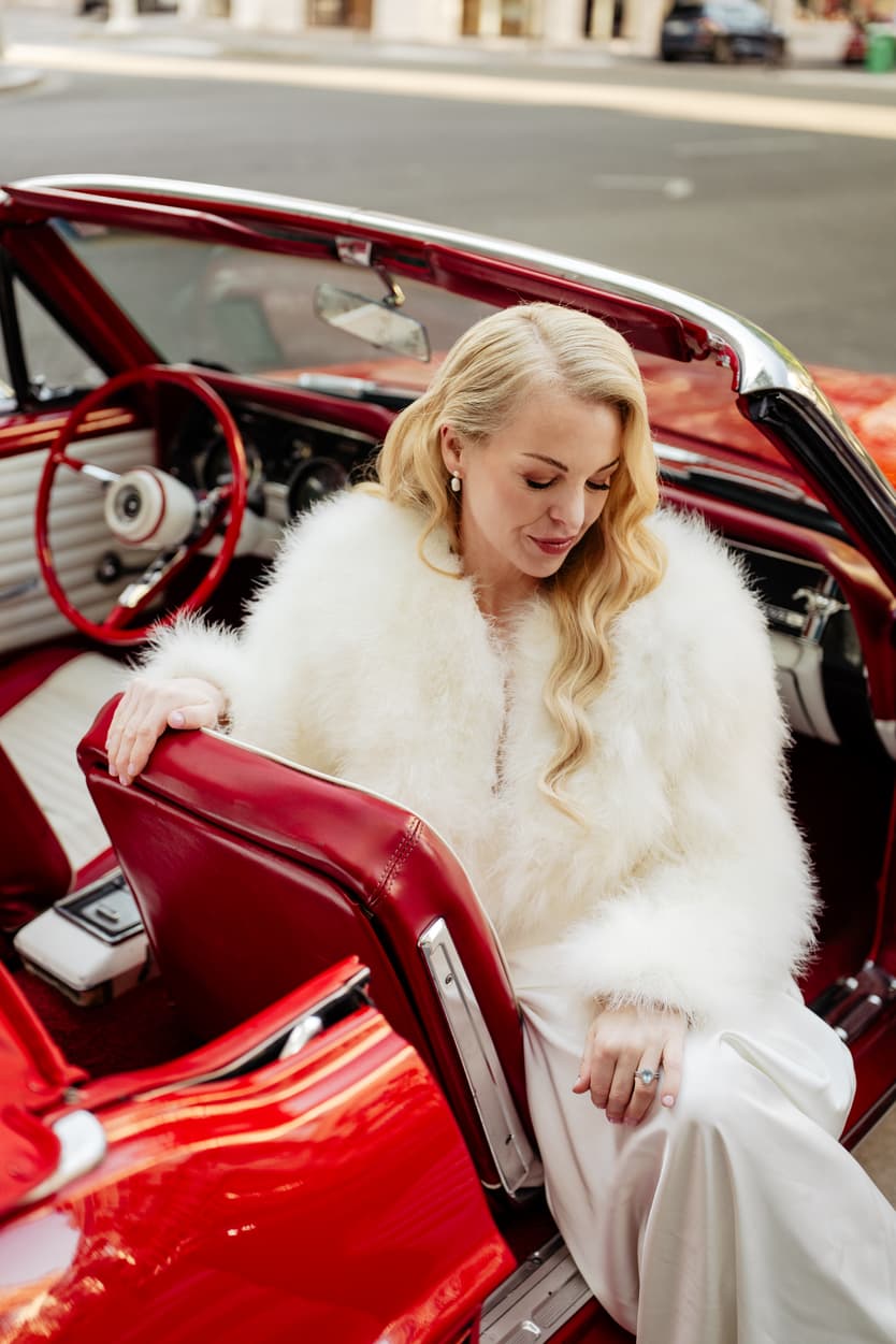 Bride in a white fur wrap sits in a red convertible on a Paris street, with the top down