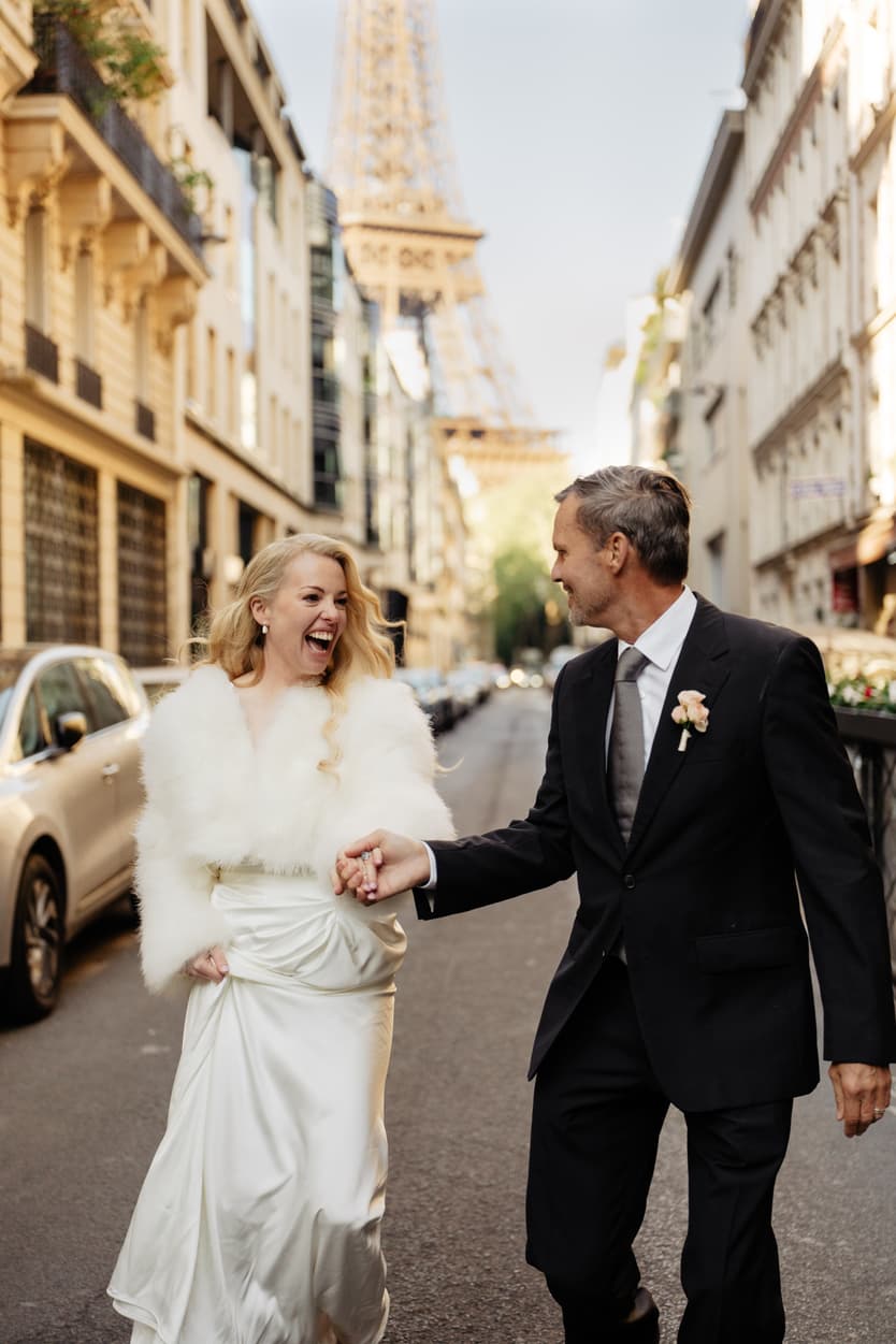 A bride laughing as she walks with her groom down a Paris street toward the Eiffel Tower
