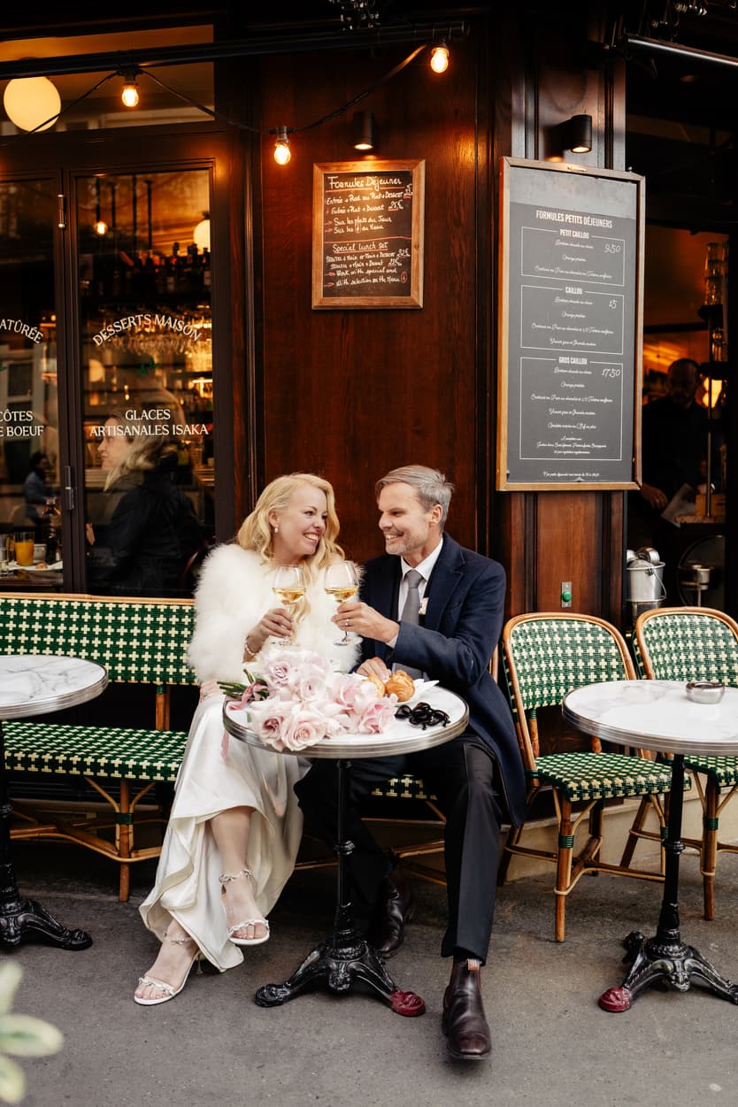 A bride and groom toast at a Paris street café in warm light, beside flowers and pastries