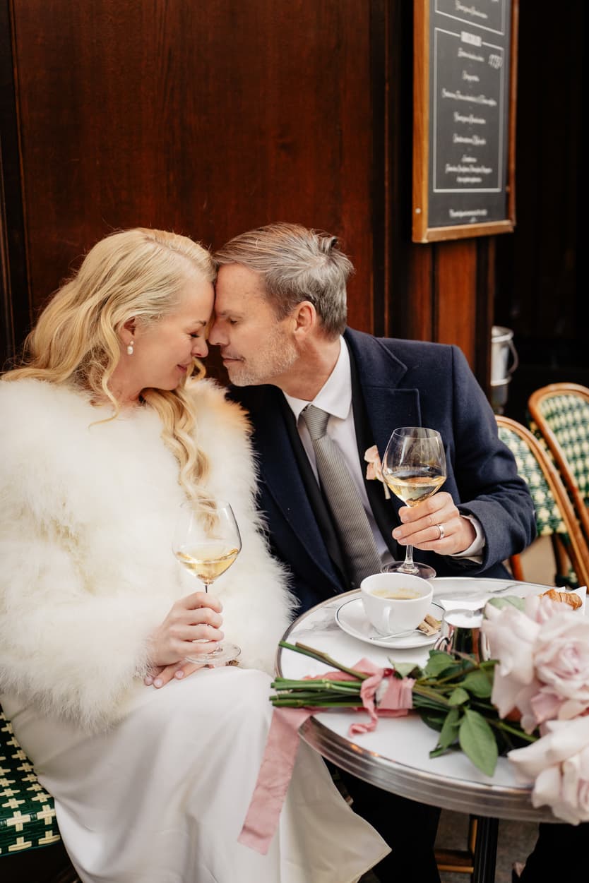 A bride and groom lean forehead to forehead at a Paris café table, holding white wine beside pink roses in warm light