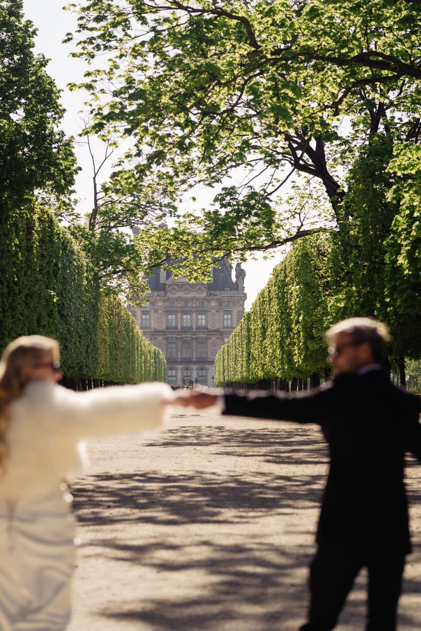 Bride and groom reach for each other in a sunlit garden under leafy trees