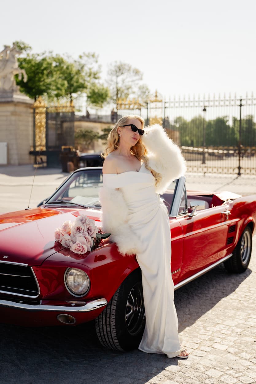 Bride in a white gown and sunglasses leans on a red convertible in bright Paris sunlight on a cobblestone street