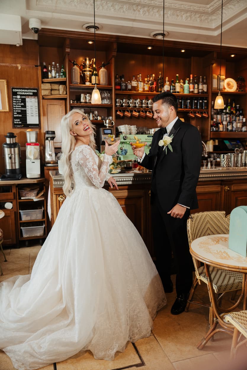 Bride and groom toast cocktails at a cozy Paris café bar with warm pendant lights above