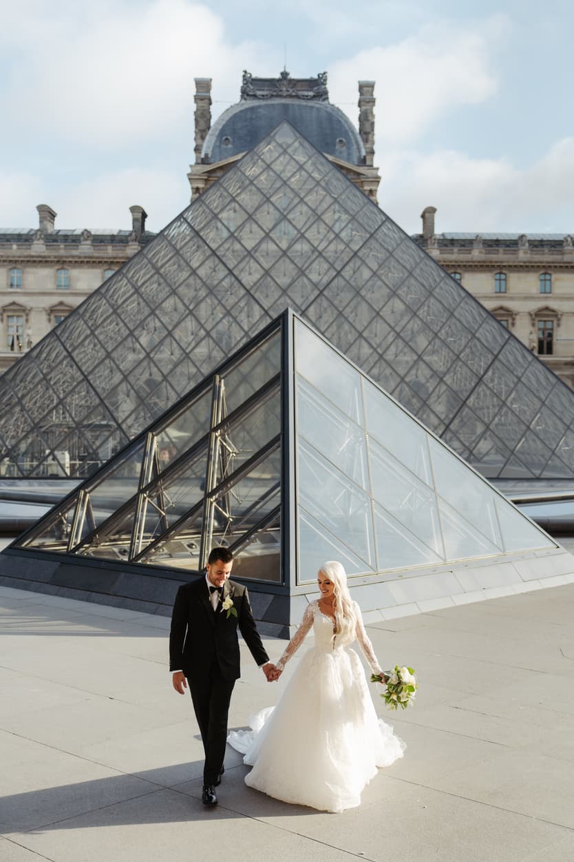 Bride and groom walk hand in hand by the Louvre pyramid in soft daylight, bouquet in her hand