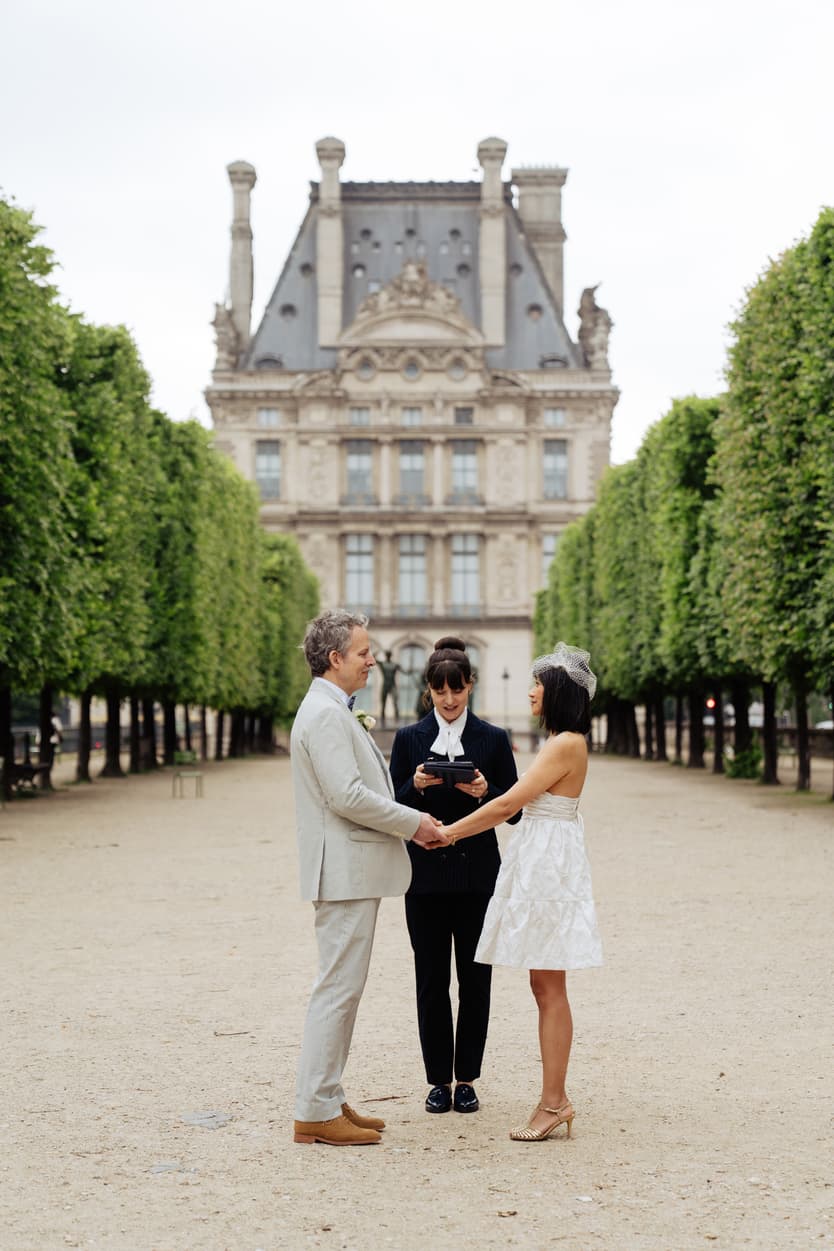 Bride and groom hold hands with an officiant in a Paris garden, framed by trees and soft daylight