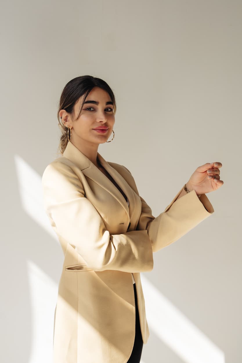 Woman in a beige blazer stands by a sunlit wall with soft diagonal light