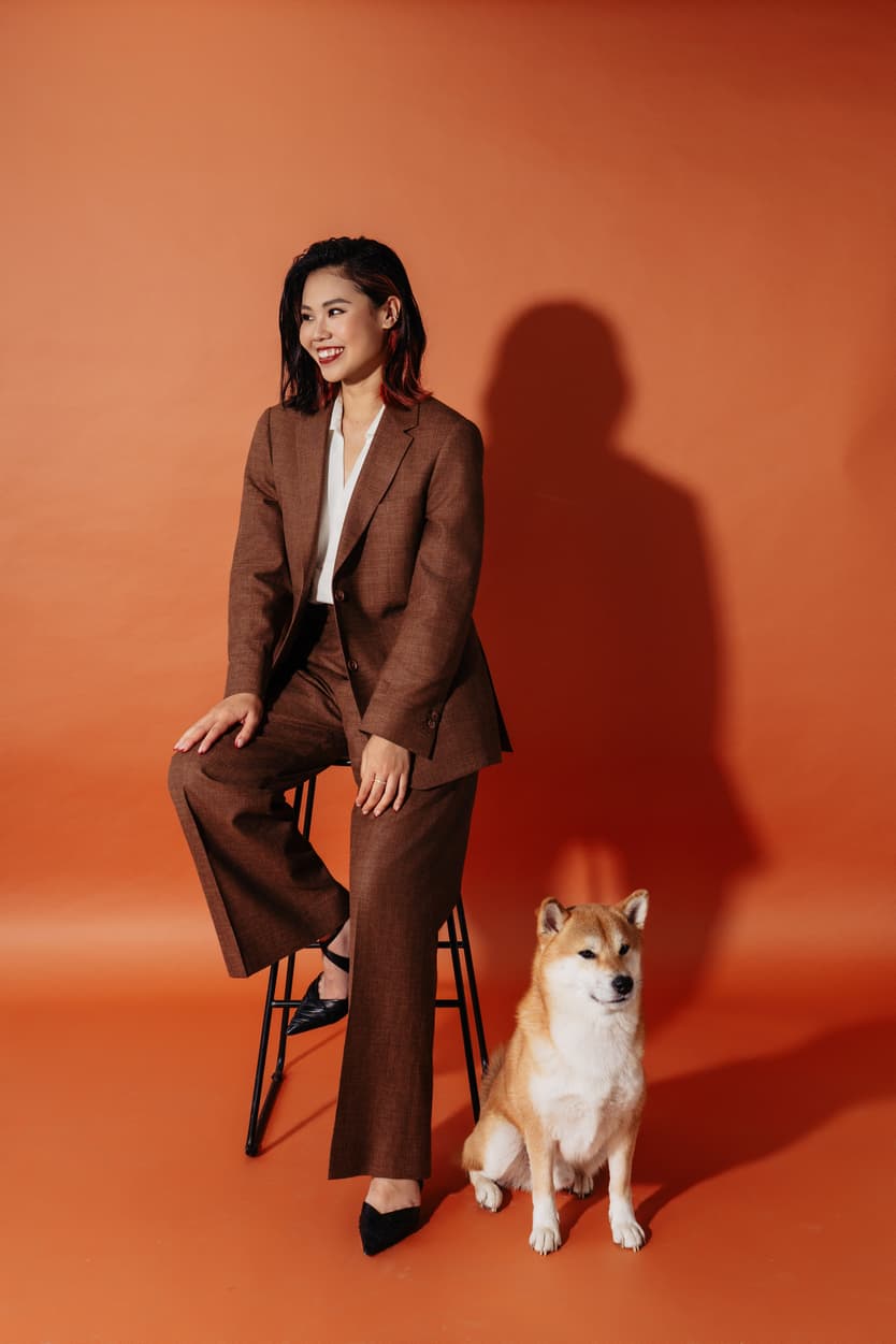 Woman in a brown suit sits on a stool beside a Shiba Inu against an orange studio backdrop