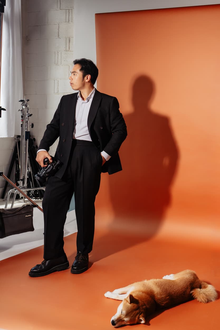 Man in a black suit holds a camera in a studio with an orange backdrop and a dog lying at his feet