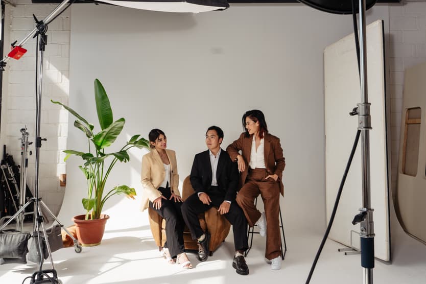 A couple sits in a bright studio beside a potted plant, framed by lighting gear