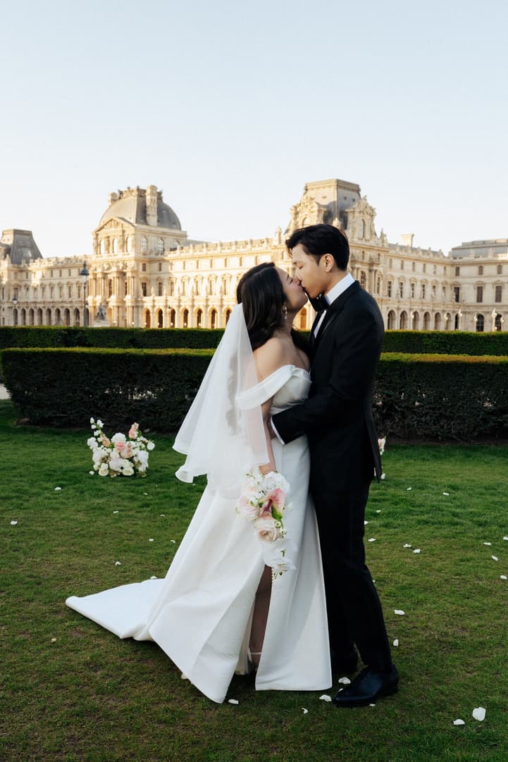 Jason kissing Nancy, her veil and bouquet in hand, in front of the Louvre