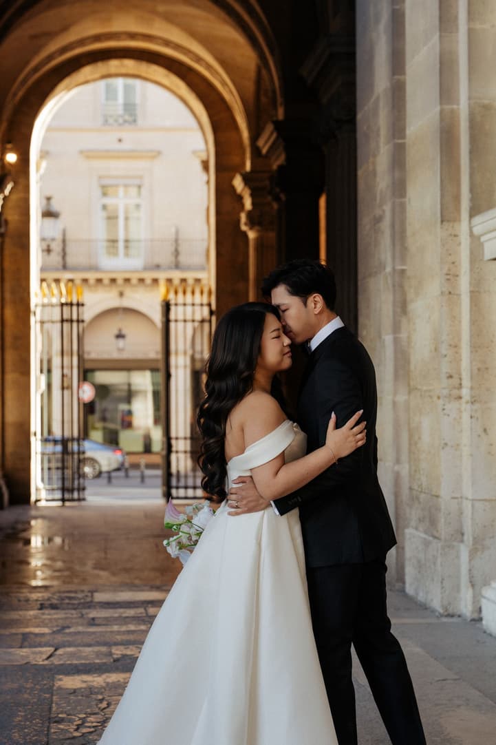 Jason kissing Nancy's forehead under an arch of the Louvre
